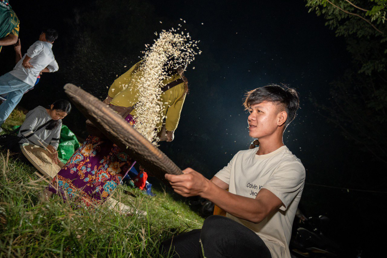 A young man winnowing rice at night, grains suspended in the air above the tray.