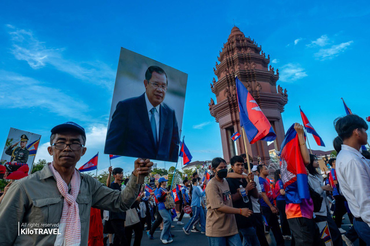 A crowd marching past the Independence Monument carrying portraits and Cambodian flags.